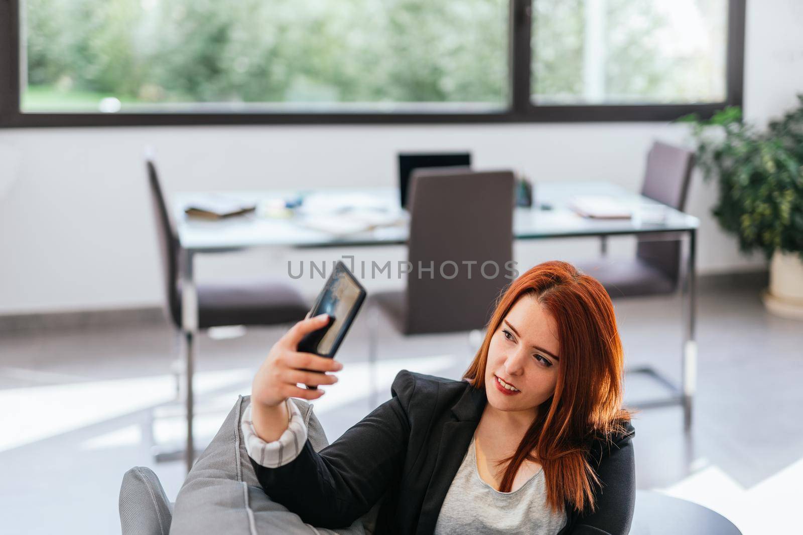 Young red-haired girl in jeans, grey T-shirt and black blazer resting on the sofa in her living room at home. She is taking a selfie with her smartphone. She has her entrepreneurial self-employed business desk behind her and large windows with natural light.