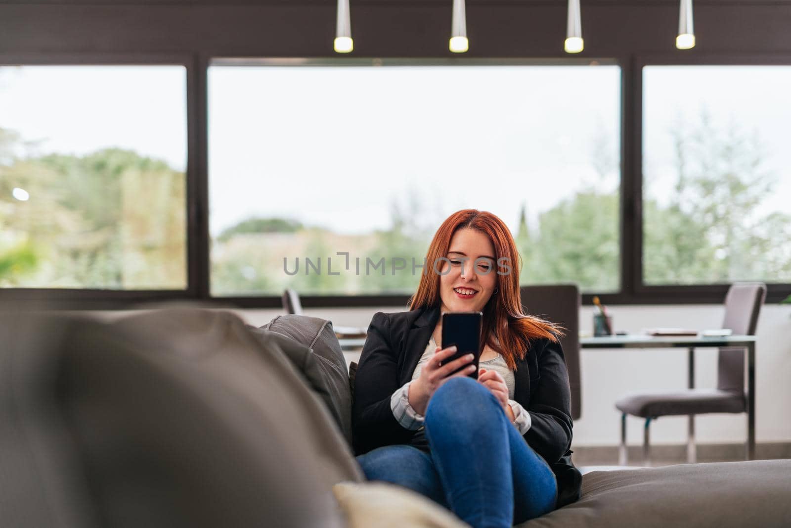 Young red-haired girl in jeans, grey T-shirt and black blazer resting on the sofa in her living room at home. She is talking on her smartphone making a video call. Large windows behind with natural light and plants.