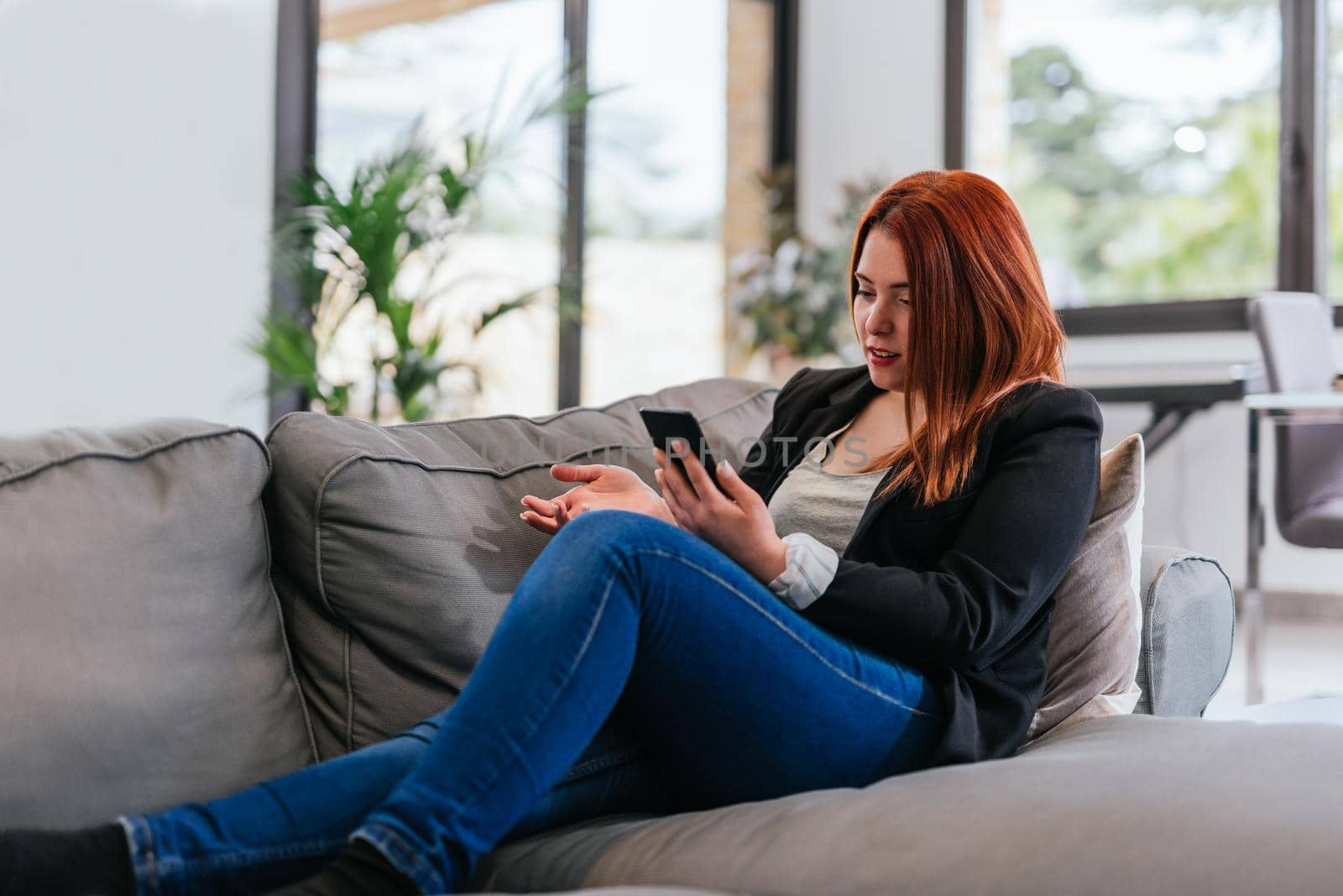 Young red-haired girl in jeans, grey T-shirt and black blazer resting on the sofa in her living room at home. She is talking on her smartphone making a video call. Large windows behind with natural light and plants.