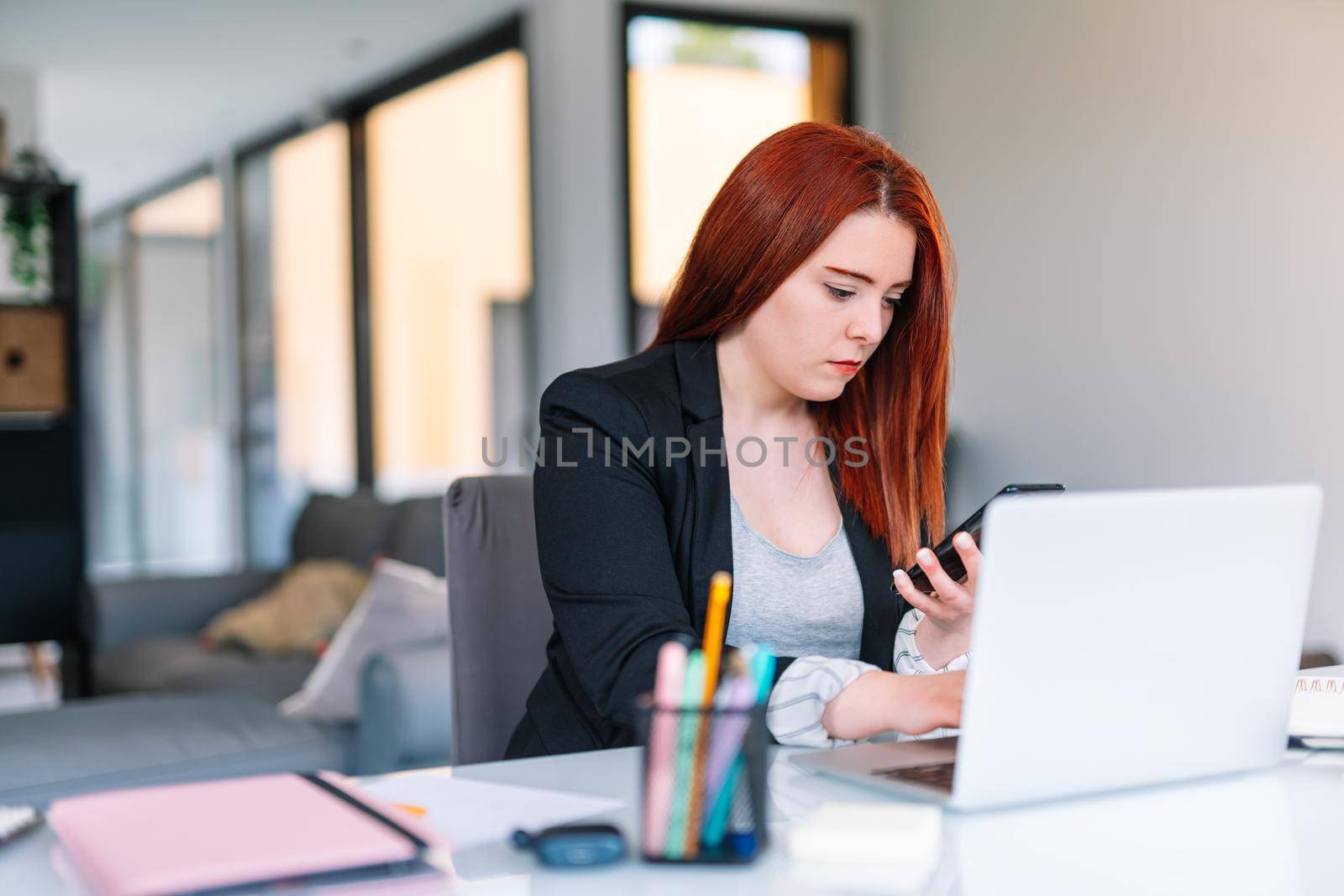 Young red-haired girl teleworking from home in her living room with her white laptop. Brightly lit scene with a large window in the background with natural light. She is wearing a black blazer. Young self-employed businesswoman and entrepreneur working at computer.