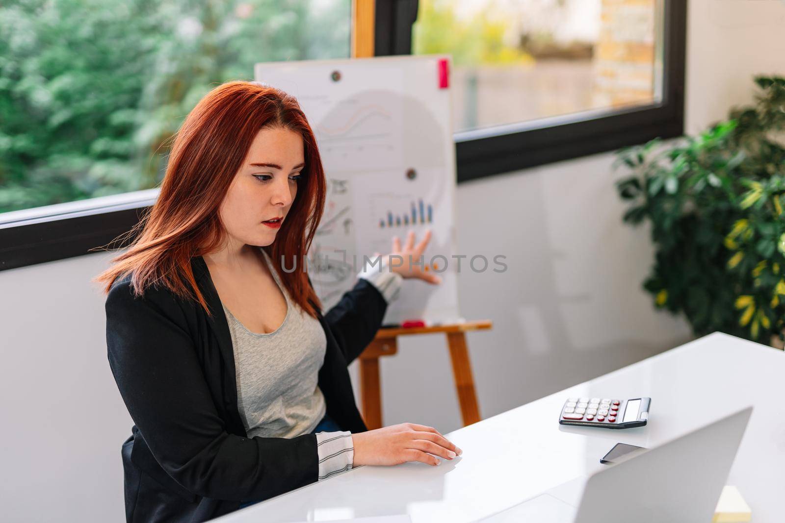 Pretty young redheaded pretty woman teleworking from home. She is making a work video call explaining something. Very bright environment with a large window behind and natural light. Young businesswoman and entrepreneur working on white laptop.