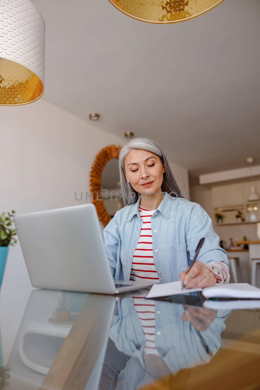 Female person sitting at the table and writing in notebook while working on modern laptop in living room