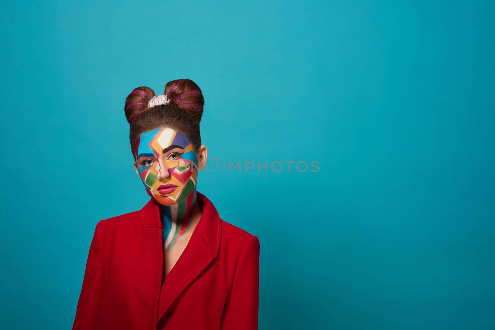 Stylish, trendy model wearing in red jacket, posing in studio with blue background. Cool girl has creative pop art make up on face and nice bow hairstyle. Funky lady looking at camera, posing.
