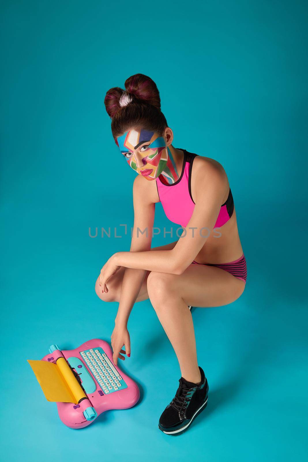 Beautiful model wearing in pink sportive top, has coroful pop art make up and bow hairstyle. Funky girl looking at camera, posing in squatting pose. Pink and blue toy typewriter.