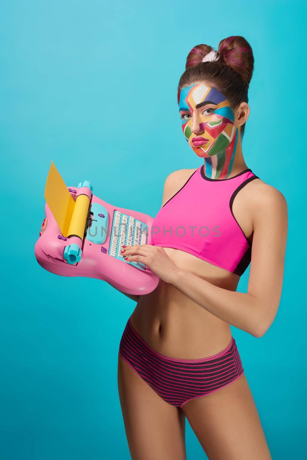 Stylish girl with slender figure looking at camera, holding pink toy typewriter, standing. Beautiful, cool model has pop art colourful make up on face, wearing pink sportive top.