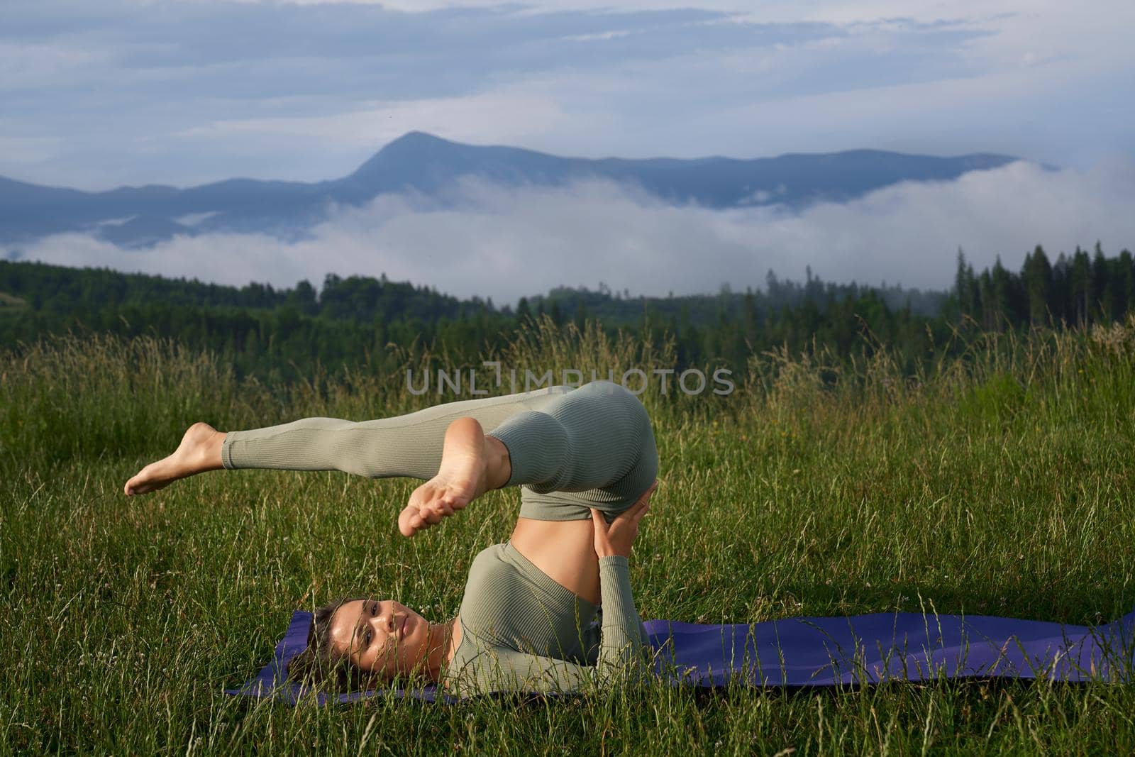 Sporty young woman in green active clothes doing acro yoga on mat among green mountains. Pretty lady taking care of body by regular workout in fresh air.