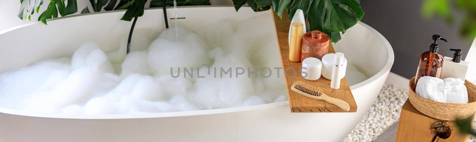 Modern bathroom interior with white bubble filled bathtub and monstera tropical plant. Eco body care products on wooden home decor elements. Concept of organic cosmetics, spa