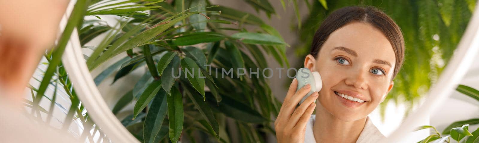 Portrait of young woman joyfully looking at herself in the mirror while cleansing her skin using silicone face brush in the bathroom decorated with green plants. Beauty, skincare, spa concept