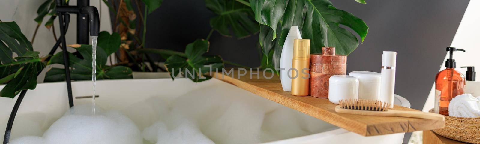 Wooden shelf for beauty and body care products on wooden shelf over modern white bubble filled bathtub. Cozy bathroom decorated with green tropical plant Monstera. Concept of organic cosmetics, spa