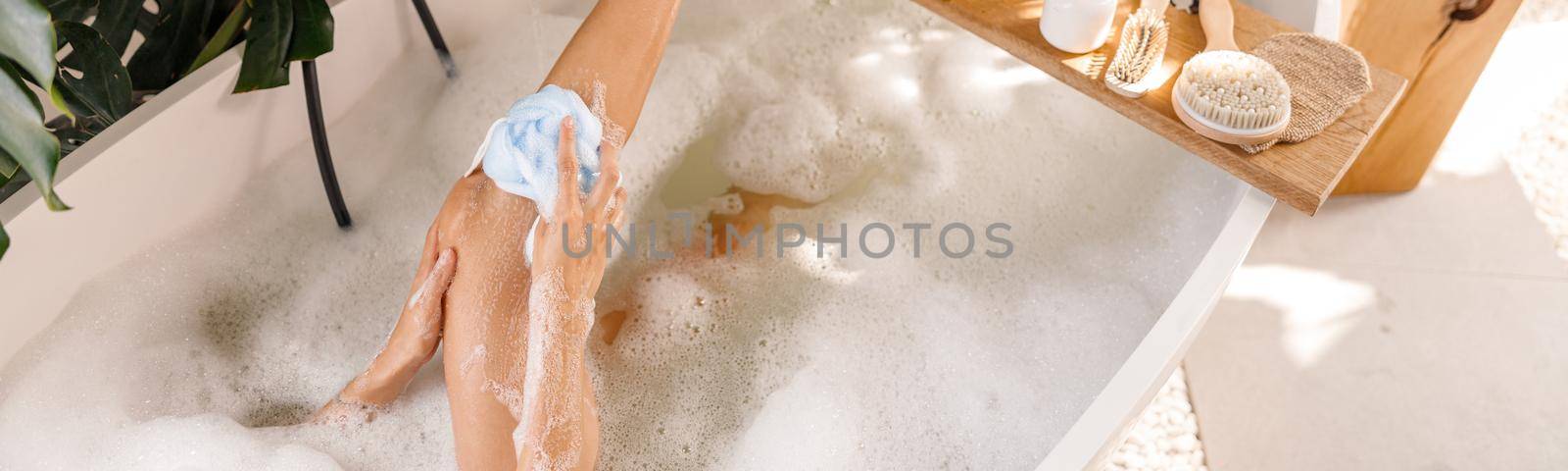 High angle view of young woman using sponge while bathing, relaxing in bubble filled bathtub. Wellness, spa resort concept