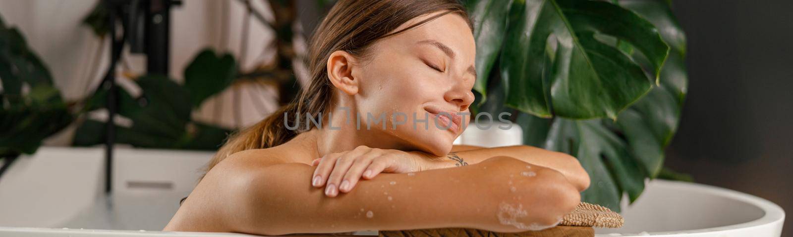 Dreamy young woman leaning on bathtub side and resting in the bathroom decorated with tropical plant. Wellness, beauty and care concept