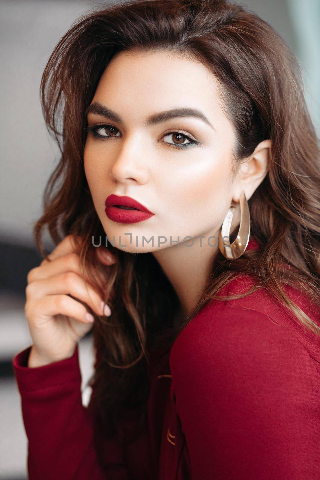 Portrait of gorgeous young brunette woman with make up, red lips, golden earrings wearing red blouse. She is looking at camera.