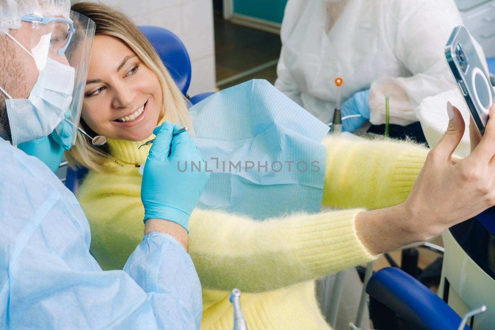 a dentist in a protective mask sits next to a patient and takes a selfie photo while working by Lobachad