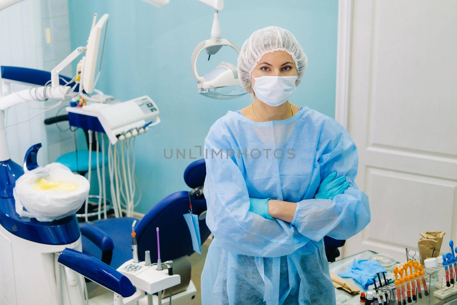 a female dentist wearing a medical mask and rubber gloves poses for the camera and folds her arms in her office.
