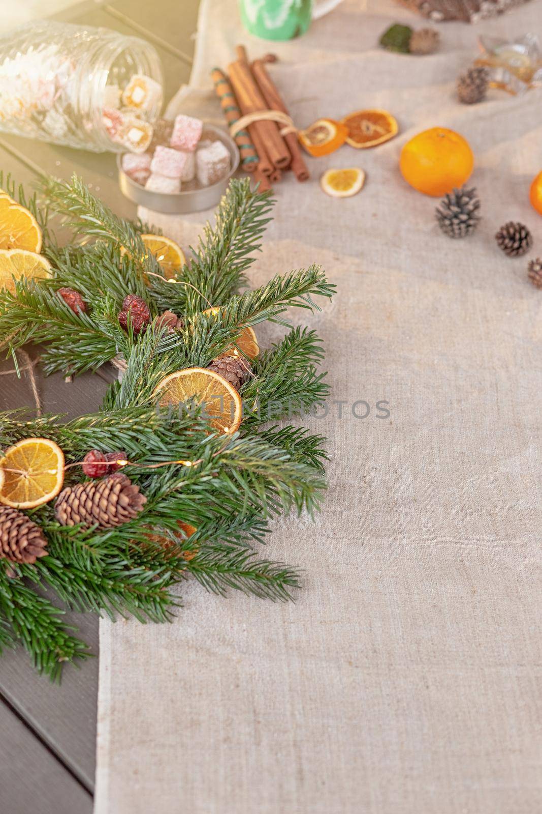 Rustic Christmas table with natural spruce wreath, dried oranges, fresh tangerines, pine cones, cinnamon sprigs and sweets on a fabric tablecloth. Copy space. Vertical