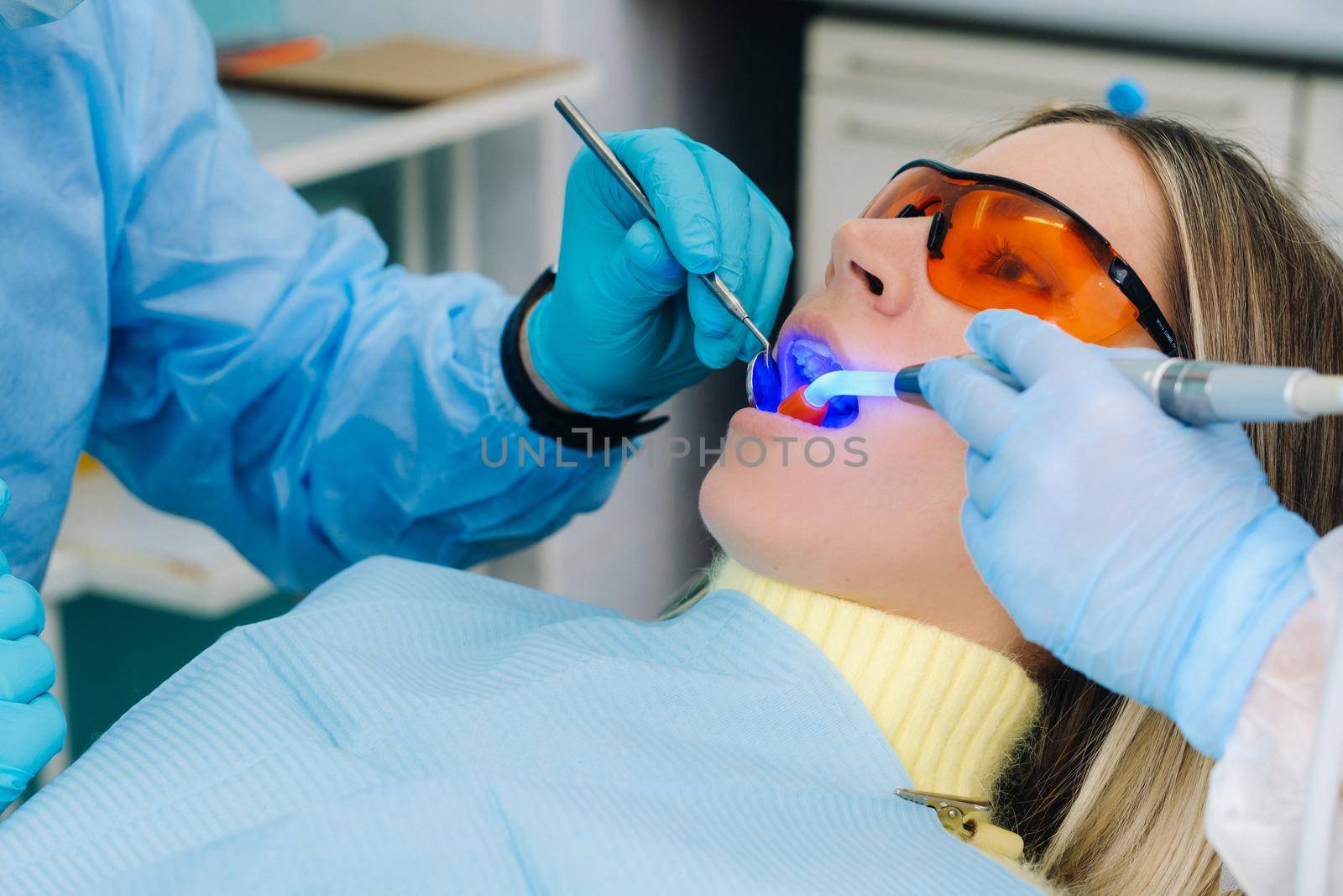A young beautiful girl in dental glasses treats her teeth at the dentist with ultraviolet light. filling of teeth by Lobachad
