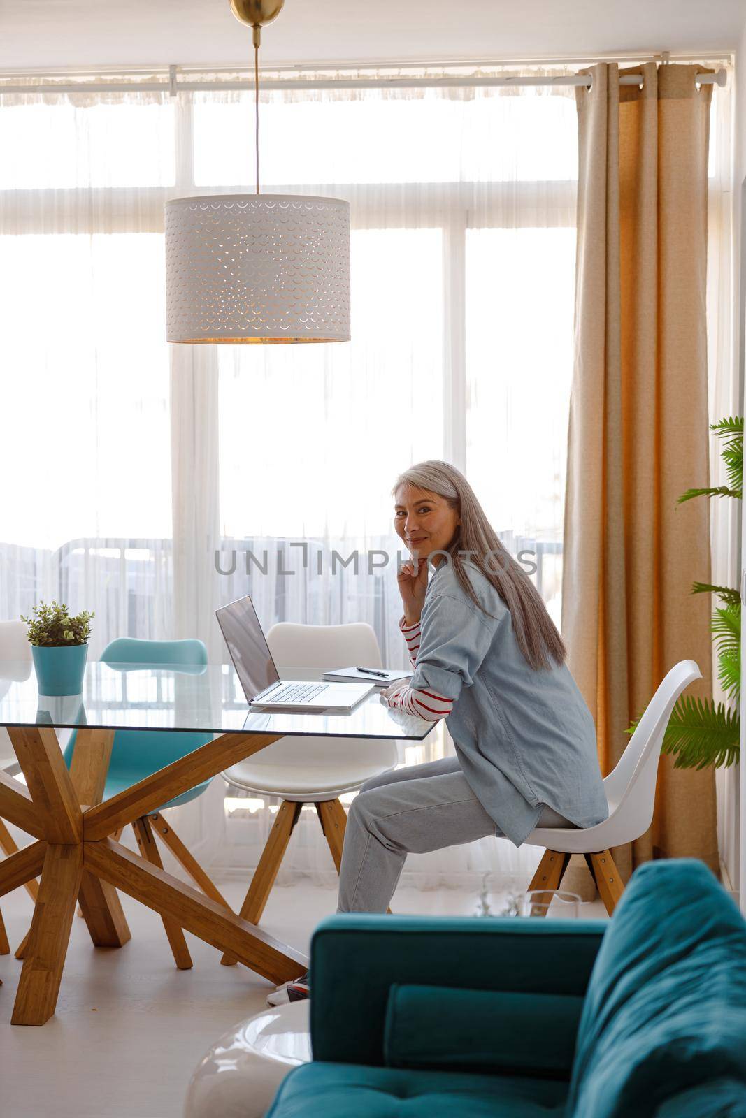 Female person looking at camera and smiling while sitting at glass desk with modern notebook at home