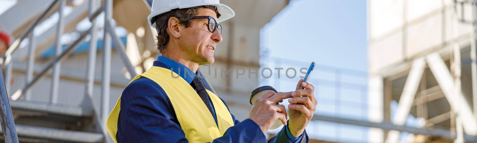 Matured man in work vest holding cup of coffee and texting message on smartphone