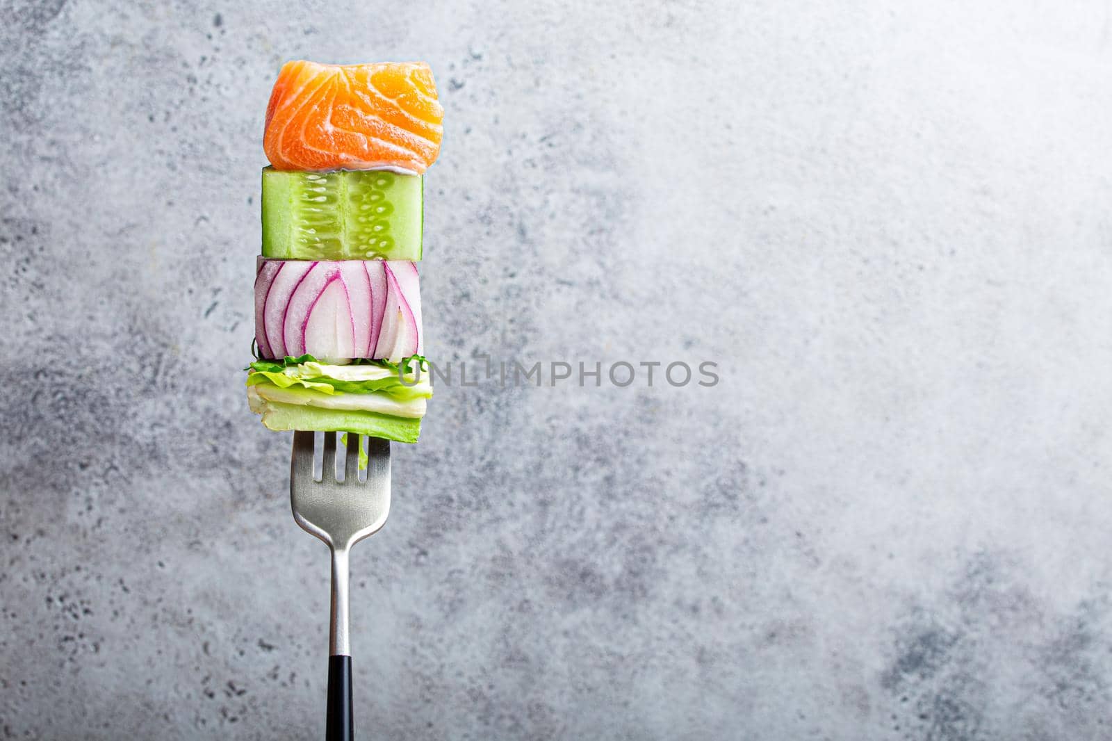 Close-up of fork with food on it: delicious fillet salmon, cucumber, onion, green salad on gray stone rustic background. Concept of healthy diet and clean eating, balanced nutrition space for text