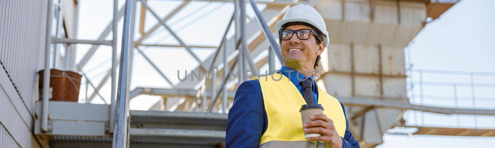 Cheerful man in work vest holding takeaway drink and folder while standing on staircase at manufacturing plant