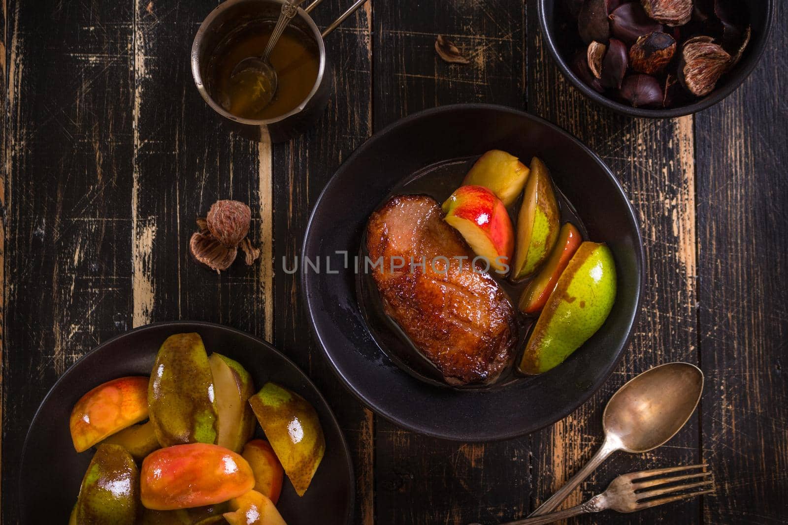 Roasted duck breast with golden crispy skin served with baked apples and chestnuts. Served on a ceramic black plate over rustic dark wooden table. Top view