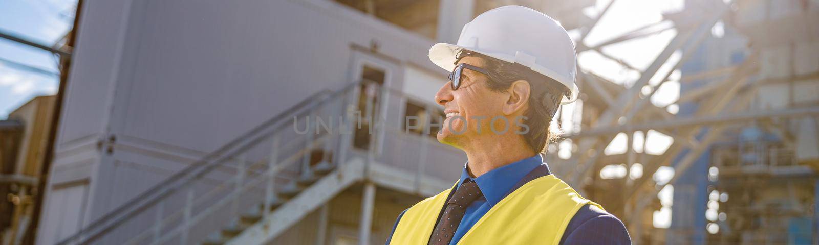 Joyful matured man holding folder with documents and smiling while standing on territory of production plant
