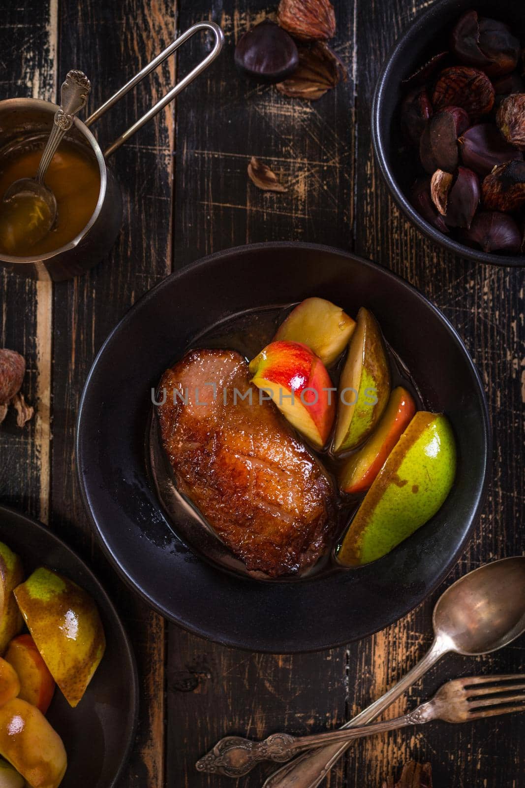 Roasted duck breast with golden crispy skin served with baked apples and chestnuts. Served on a ceramic black plate over rustic dark wooden table. Top view