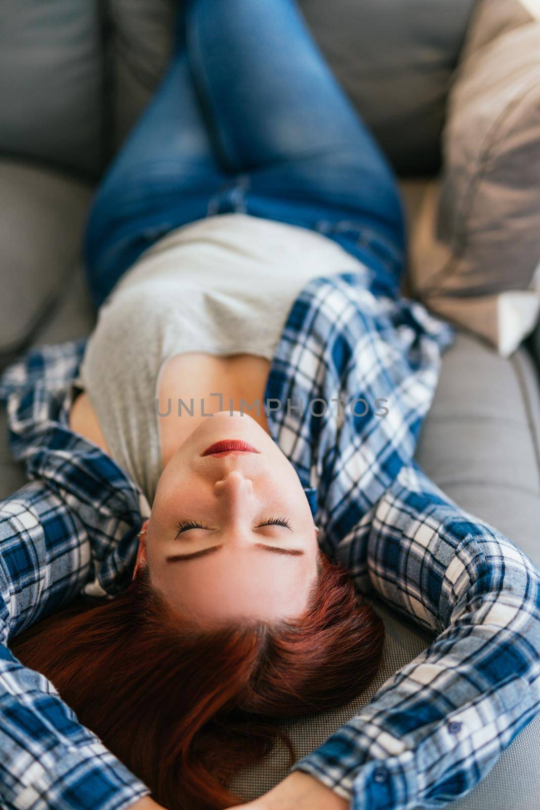 Young red-haired girl in grey T-shirt, blue and white checked shirt and jeans relaxing on the sofa. Vertical body shot while resting from a day's work.