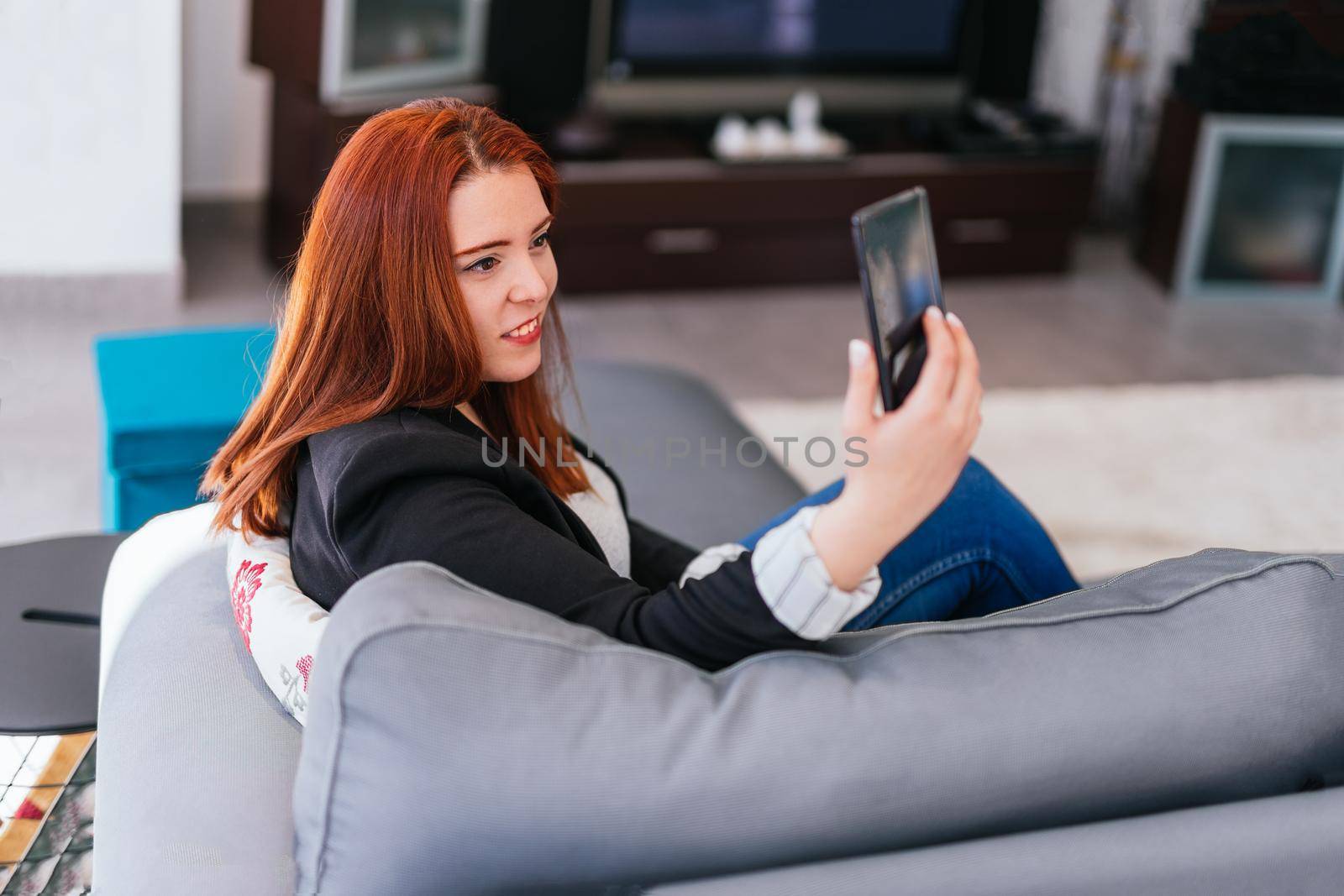 Young red-haired girl in jeans, grey T-shirt and black blazer resting on the sofa in her living room at home. She is taking a selfie with her smartphone. She has her entrepreneurial self-employed business desk behind her and large windows with natural light.