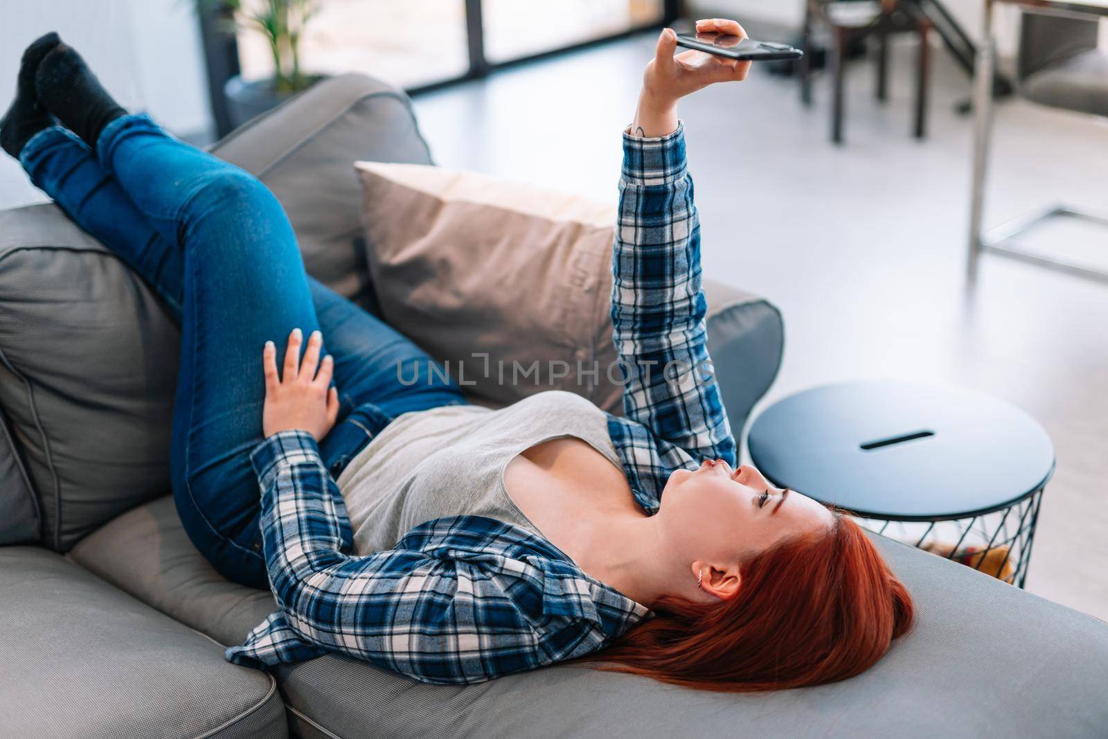 Young red-haired girl in jeans, grey T-shirt and blue shirt resting on the sofa in her living room at home. She is taking a selfie with her smartphone.