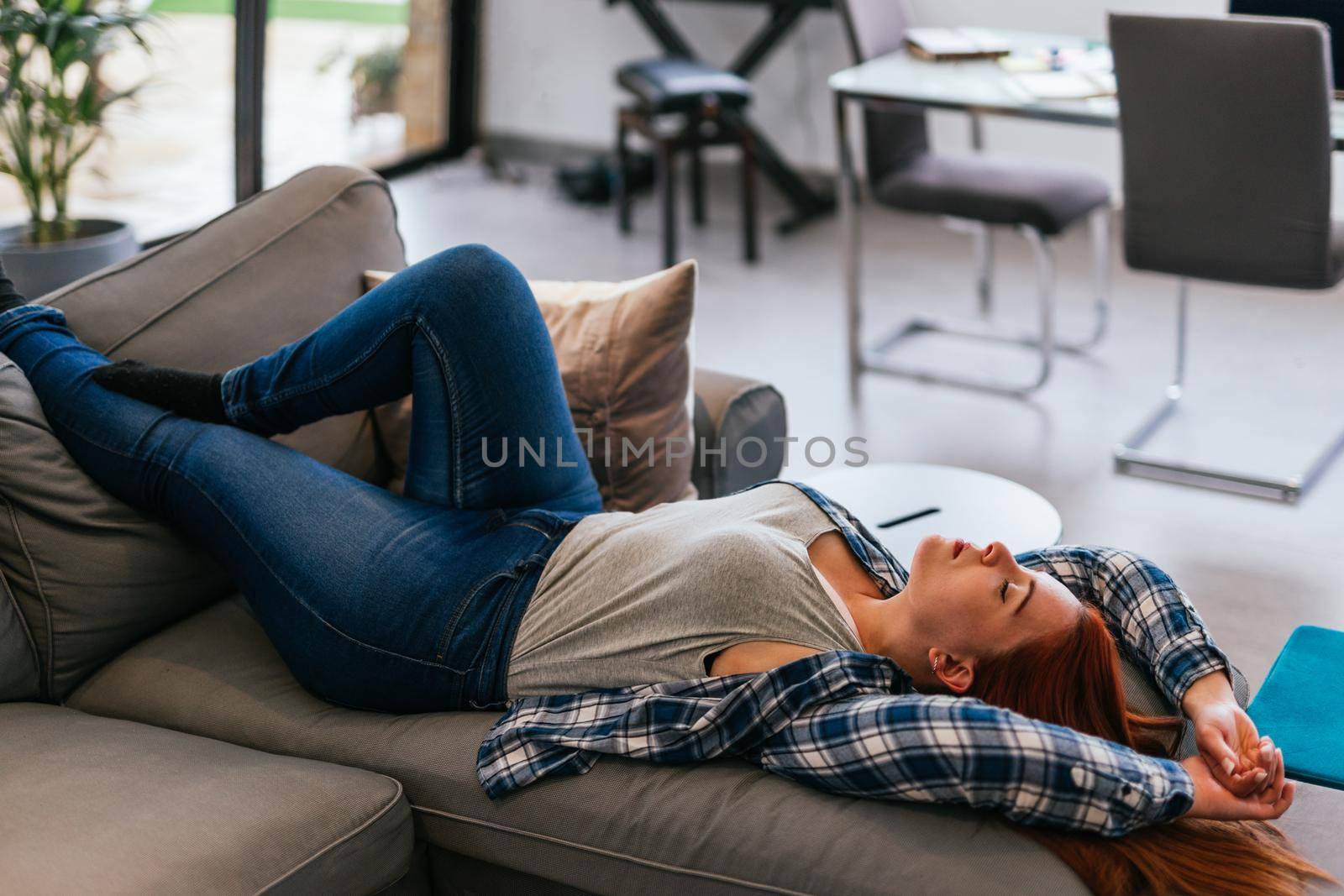 Young red-haired girl in grey T-shirt, blue and white checked shirt and jeans relaxing on the sofa. Vertical body shot while resting from a day's work.