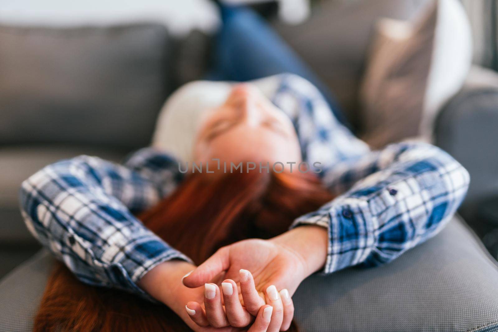 Detailed plan of the hands of young red-haired girl in grey T-shirt, blue and white checked shirt and jeans relaxing on the sofa. Vertical body shot while resting from a day's work.