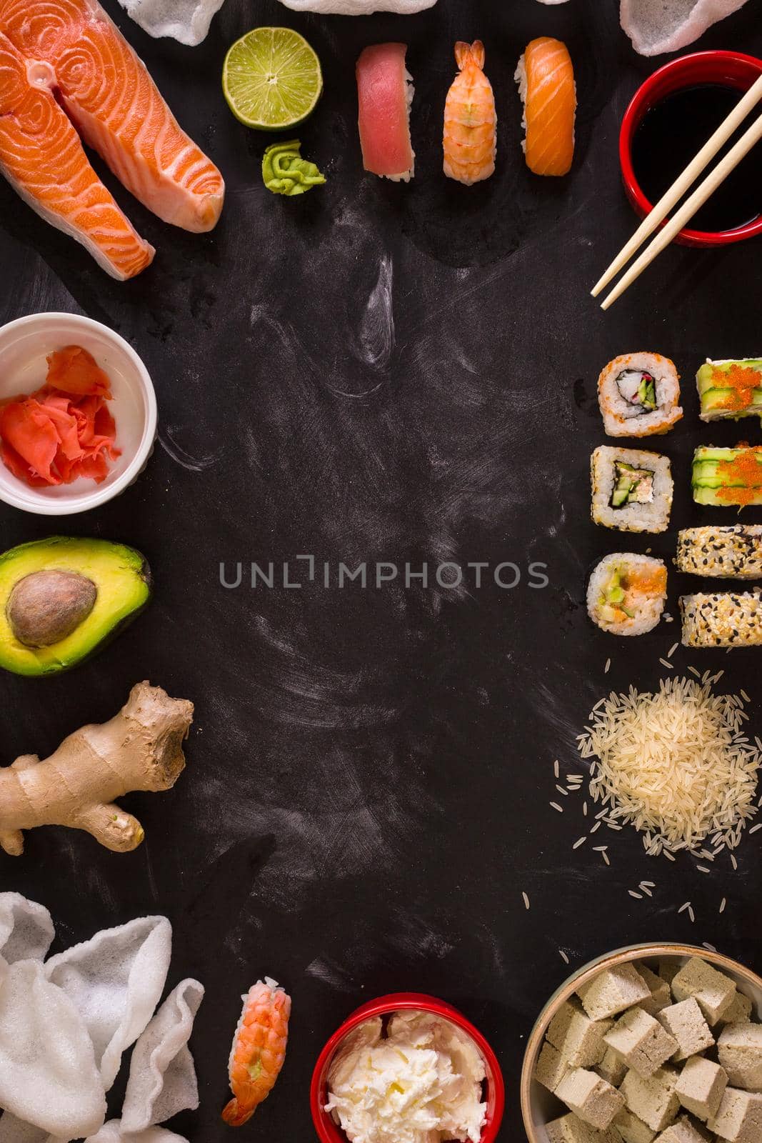 Overhead shot of sushi and ingredients on dark background. Sushi rolls, nigiri, raw salmon steak, rice, cream cheese, avocado, lime, pickled ginger (gari), raw ginger, wasabi, soy sauce, nori, сhopsticks. Asian food background. Space for text....