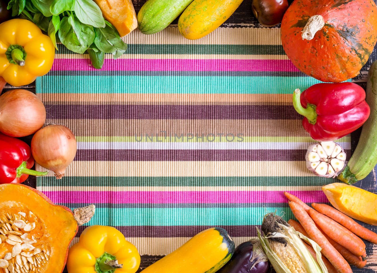 Fresh vegetables on a colorful striped kitchen towel and old rustic dark textured table. Autumn background. Healthy eating frame. Sliced pumpkin, bell peppers, carrots, onions, cut garlic, tomatoes, rucola and basil. Top view. Space for text