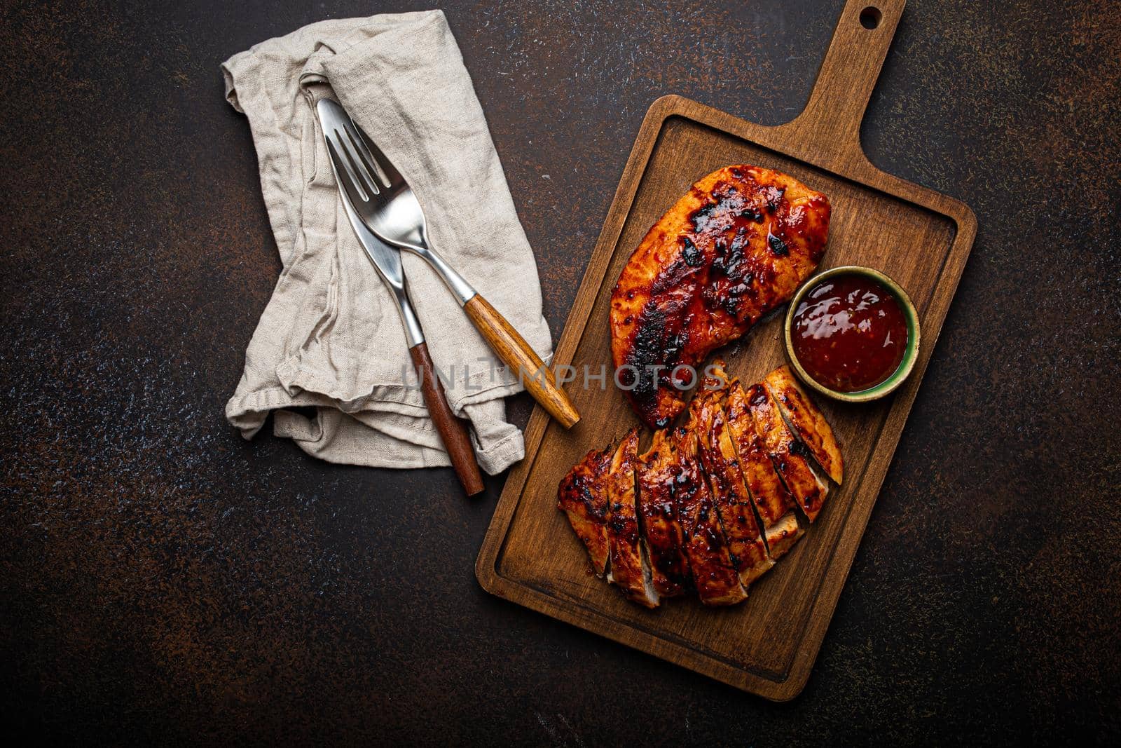 Grilled turkey or chicken marinated fillet with red sauce served and sliced on wooden cutting board on stone brown background from above, poultry breast barbecue