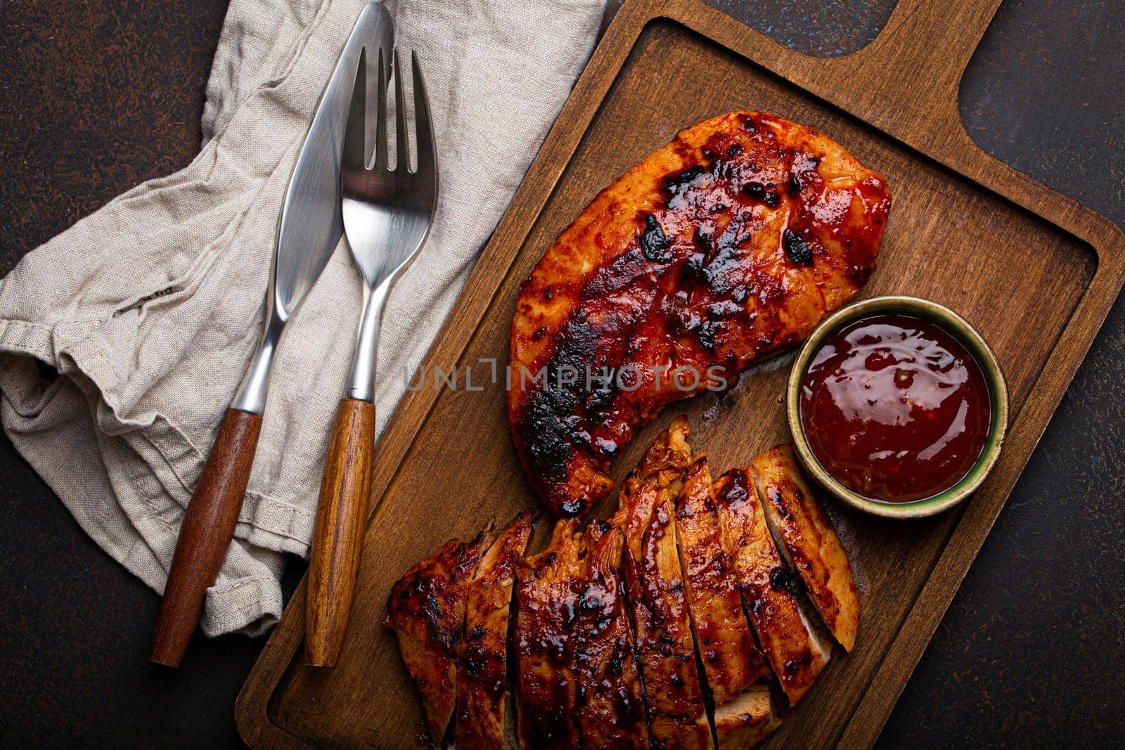 Grilled turkey or chicken marinated fillet with red sauce served and sliced on wooden cutting board on stone brown background from above, poultry breast barbecue
