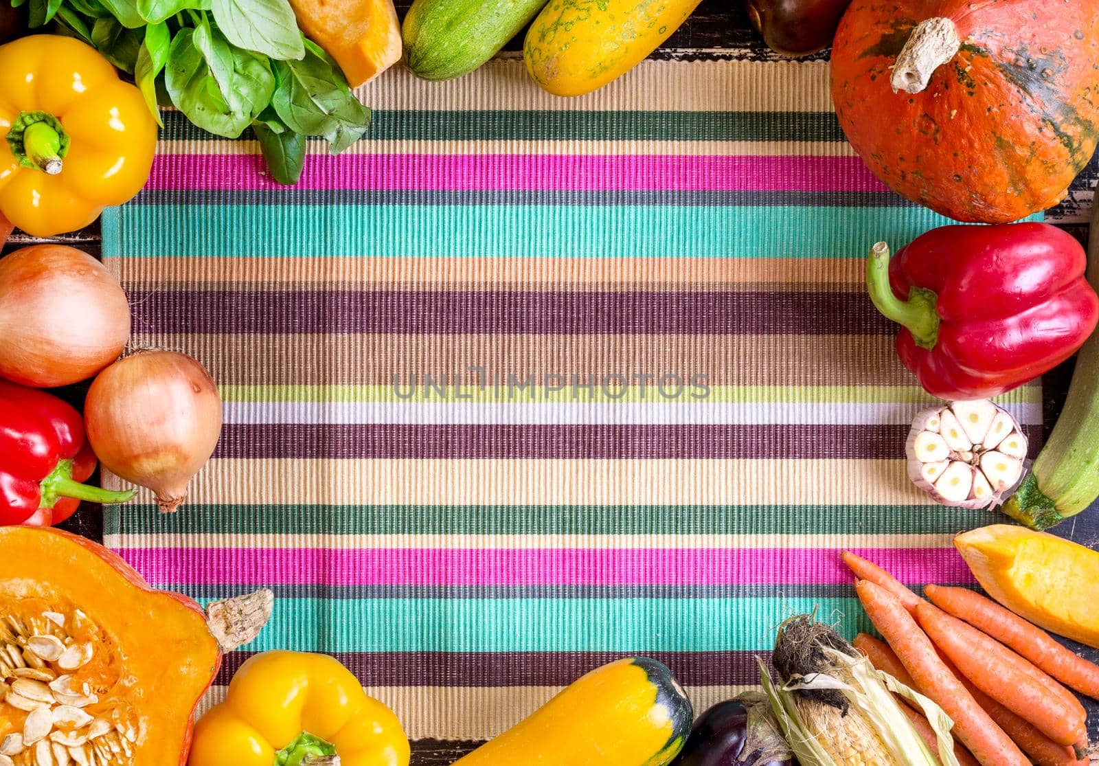 Fresh vegetables on a colorful striped kitchen towel and old rustic dark textured table. Autumn background. Healthy eating frame. Sliced pumpkin, bell peppers, carrots, onions, cut garlic, tomatoes, rucola and basil. Top view. Space for text