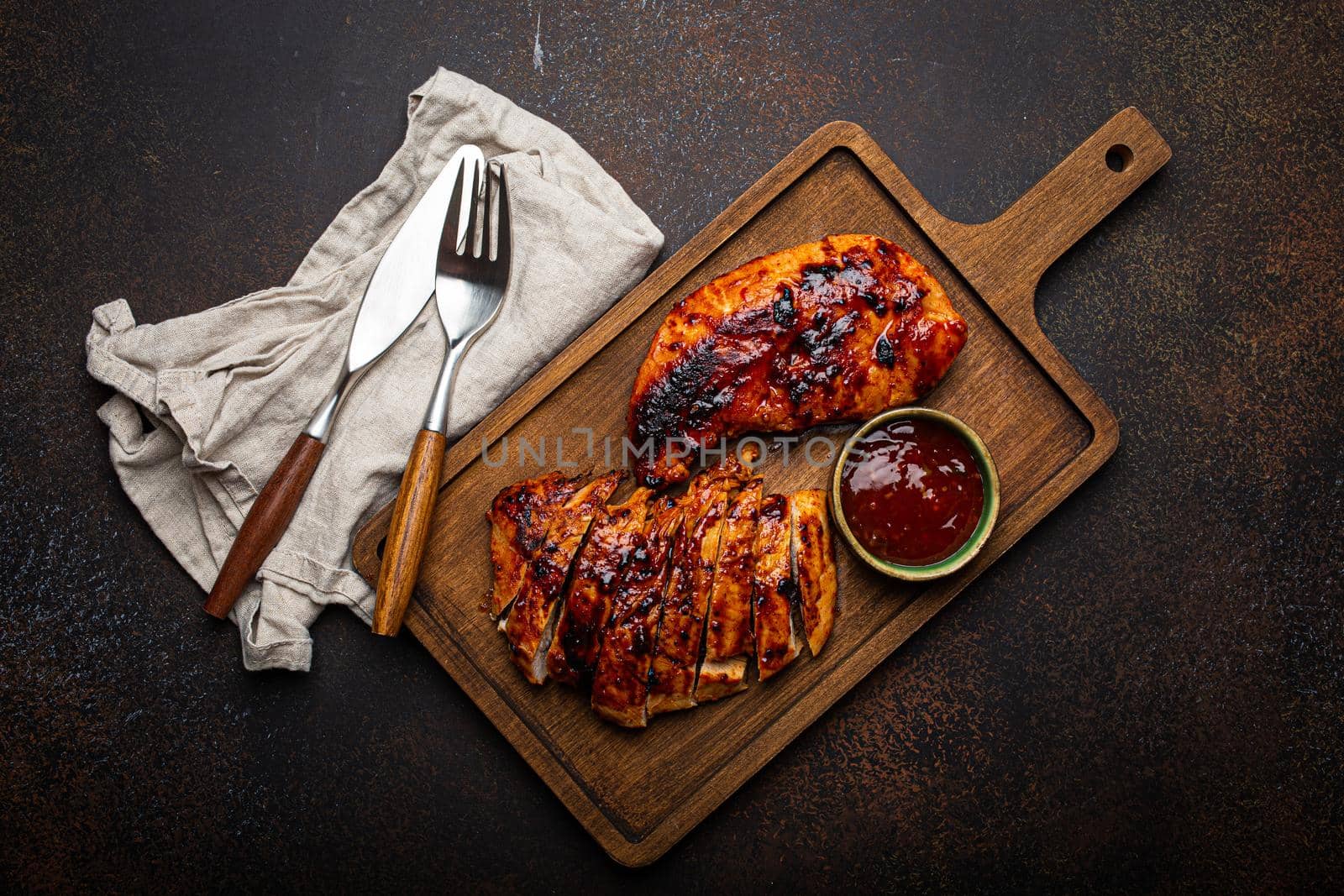 Grilled turkey or chicken marinated fillet with red sauce served and sliced on wooden cutting board on stone brown background from above, poultry breast barbecue