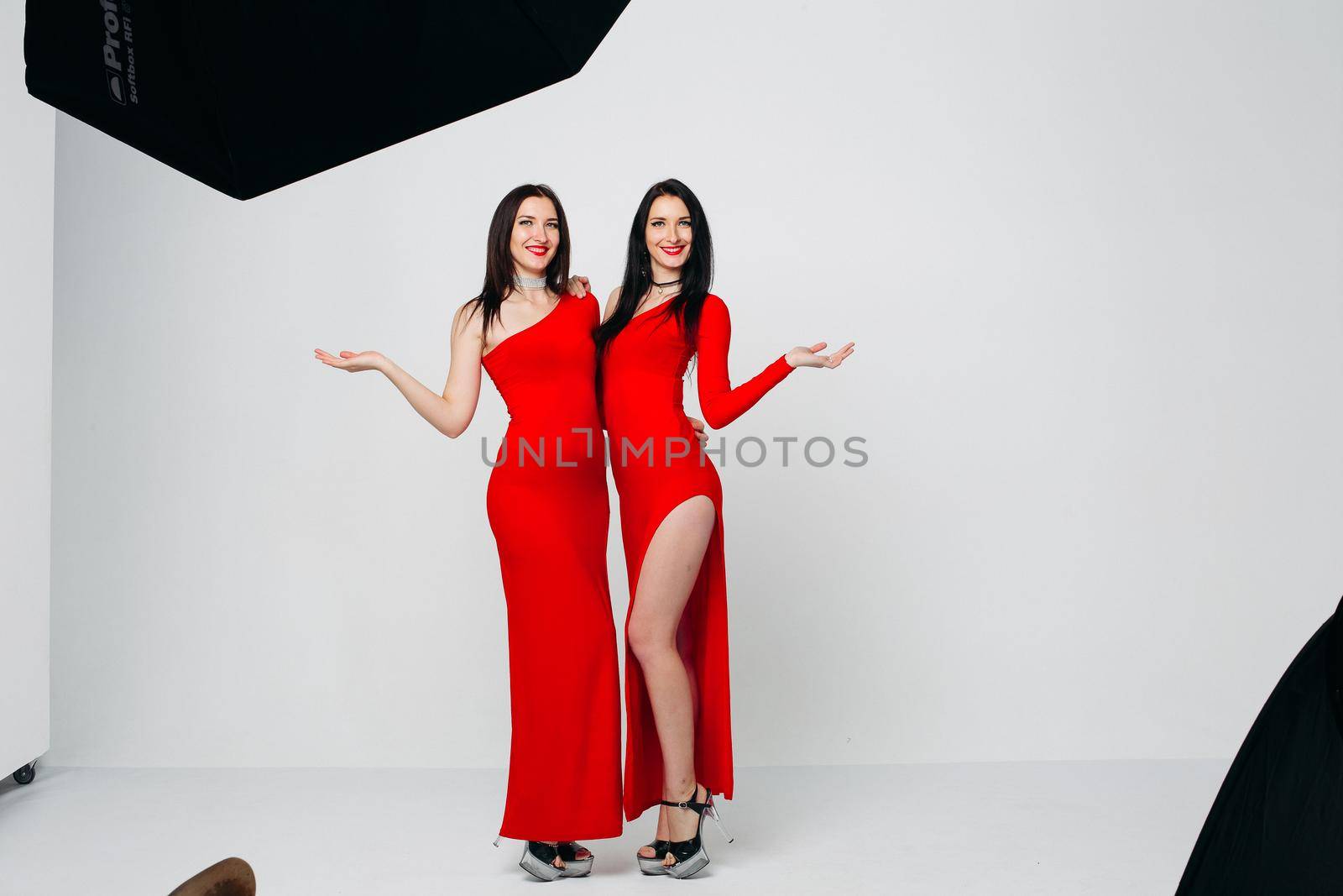 Two sexy and beautiful sisters twins in red dresses posing, looking at camera holding hands up. Pretty dancing ladies with long hair. Fashionable and stylish girls. Shopping, fashion, party.