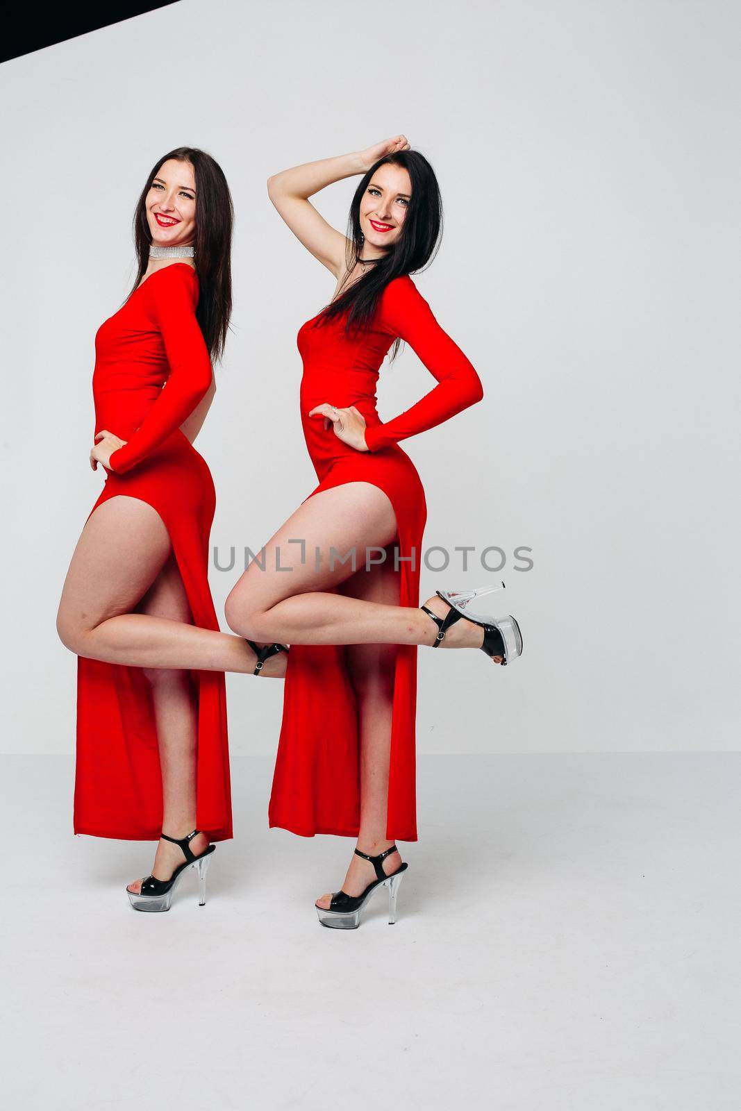 Two sexy and beautiful sisters twins in red dresses posing, looking at camera holding hands up. Pretty dancing ladies with long hair. Fashionable and stylish girls. Shopping, fashion, party.