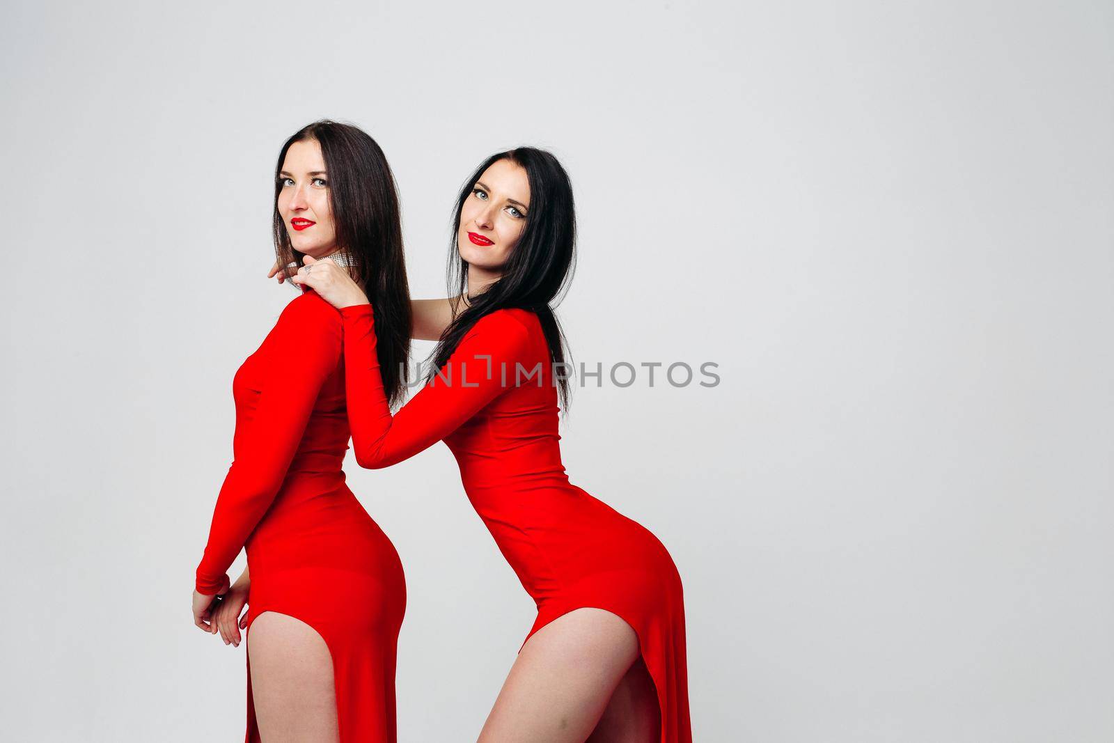 Two sexy and beautiful sisters twins in red dresses posing, looking at camera holding hands up. Pretty dancing ladies with long hair. Fashionable and stylish girls. Shopping, fashion, party.