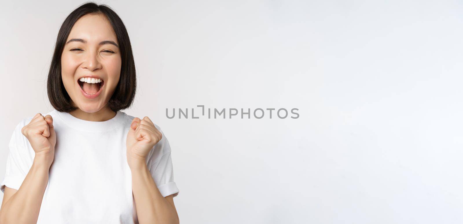 Portrait of enthusiastic asian woman winning, celebrating and triumphing, raising hands up, achieve goal or success, standing over white background.