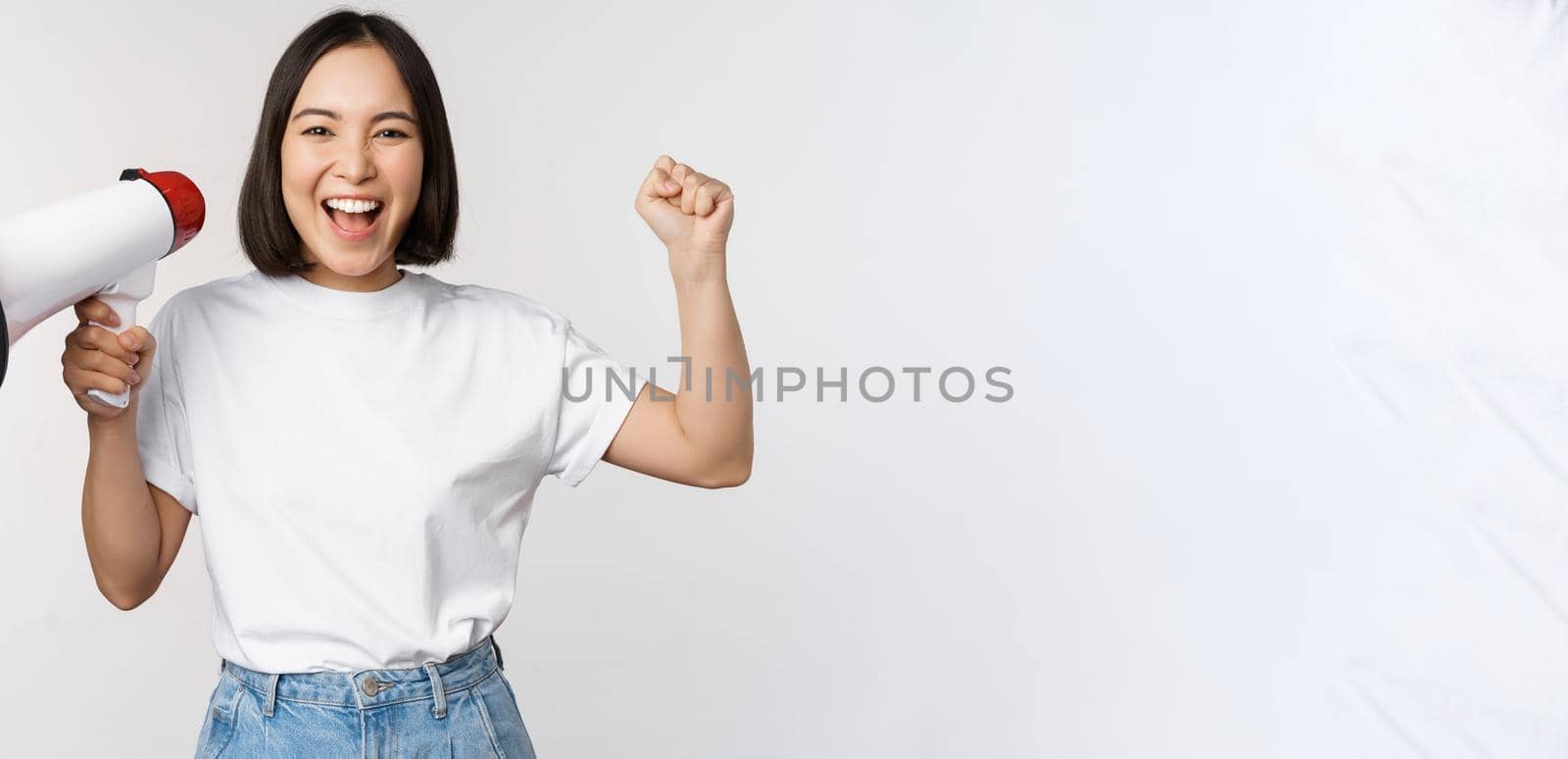 Happy asian woman shouting at megaphone, making announcement, advertising something, standing over white background by Benzoix