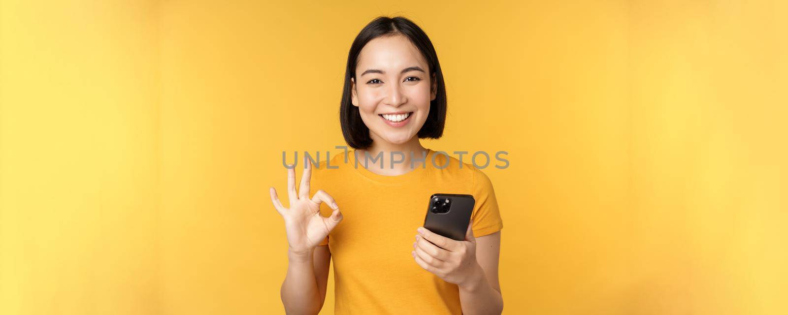 Happy smiling asian girl holding mobile phone and showing okay, recommending application on smartphone, standing over yellow background.