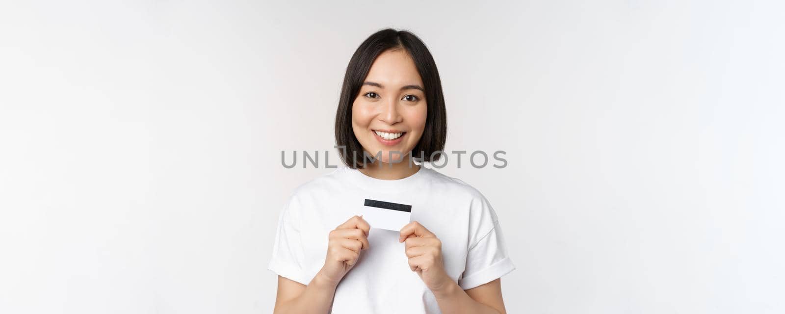 Smiling korean woman showing credit card with happy face, standing in tshirt over white background by Benzoix