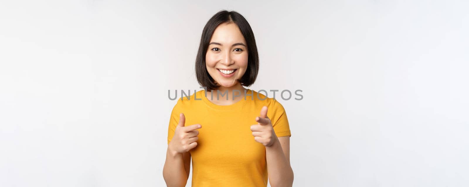 Young korean girl student, pointing fingers at camera and smiling, congratulating you, choosing, inviting people, standing over white background.