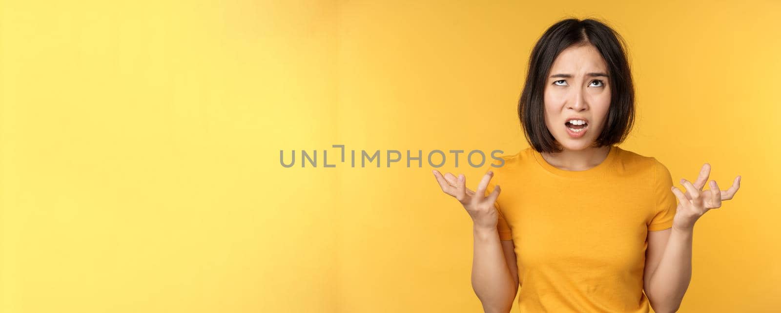 Image of angry asian woman, shouting and cursing, looking outraged, furious face expression, standing over yellow background.