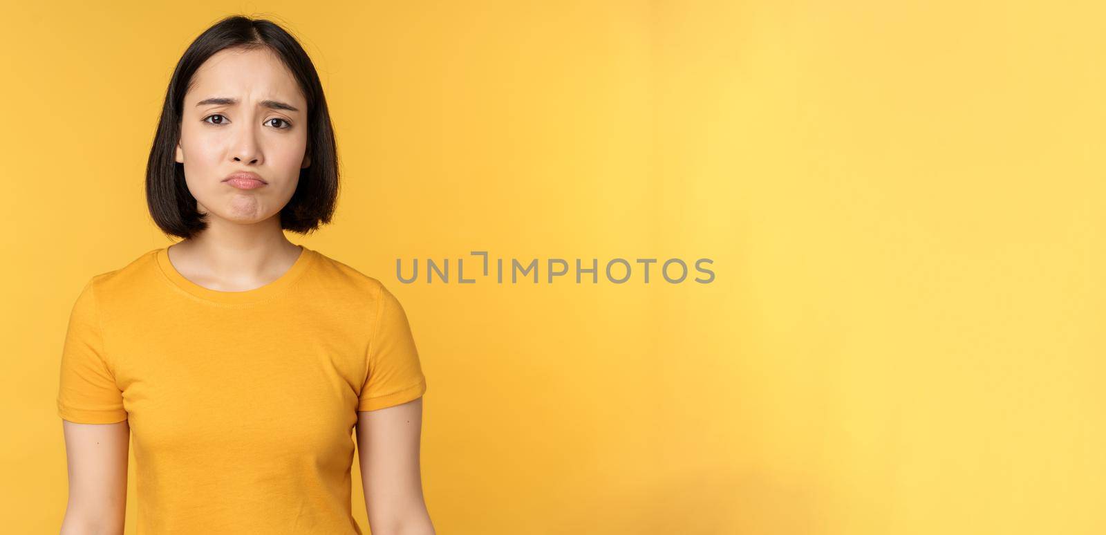Disappointed asian girl sulking, looking upset, feel unair, standing in yellow t-shirt over white background by Benzoix