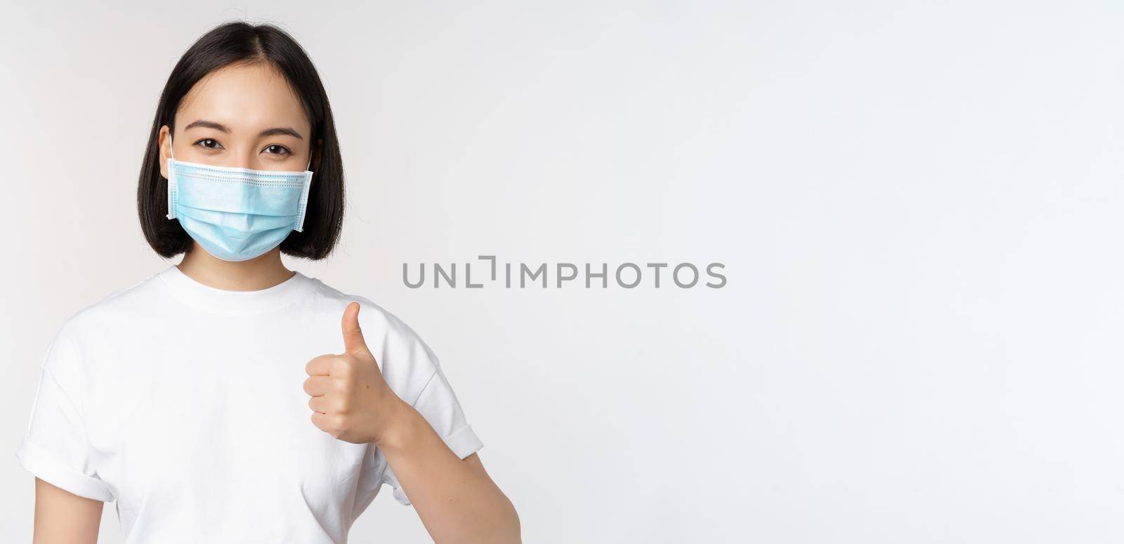 Smiling asian girl in medical mask showing thumbs up, approve something good, praise and compliment company, standing over white background by Benzoix