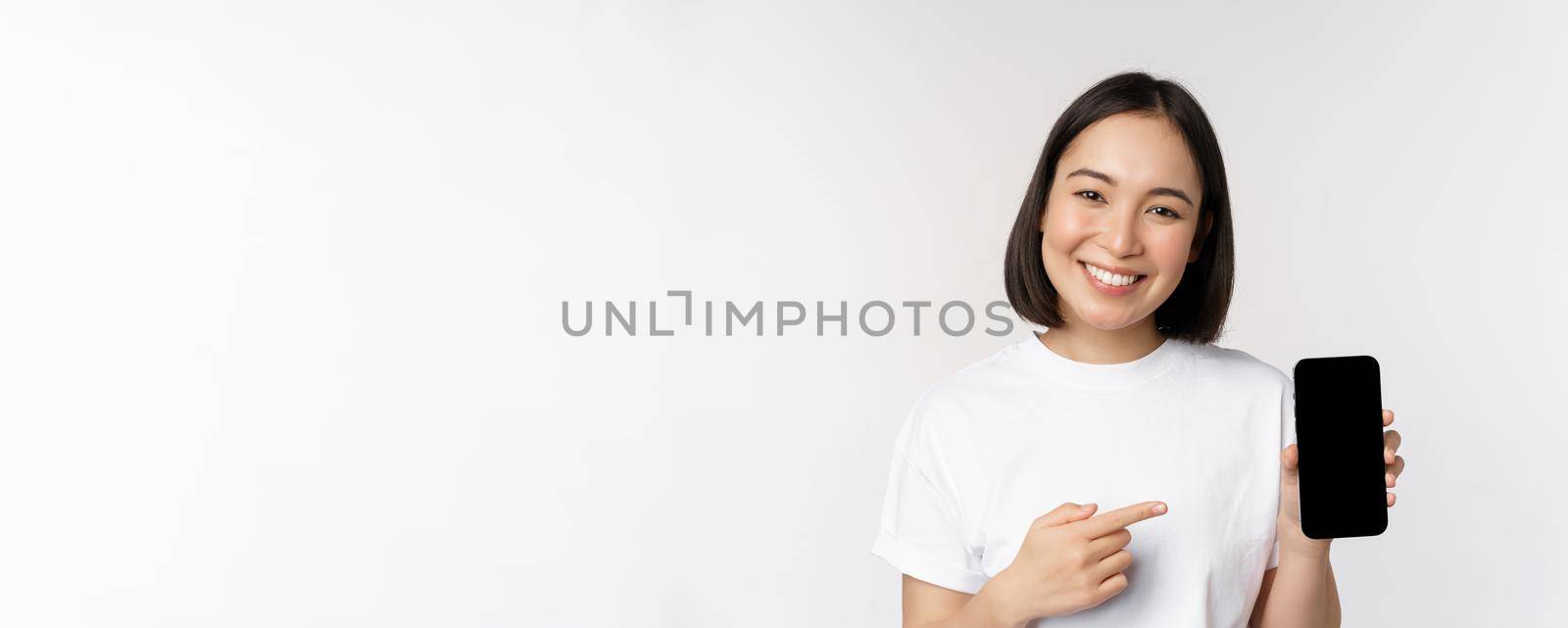 Smiling asian woman pointing finger at smartphone screen, showing application interface, mobile phone website, standing over white background by Benzoix